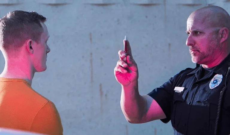 A police officer in uniform is holding a penlight, illuminated by pink lighting, against a concrete backdrop.