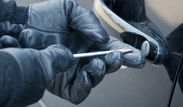 A gloved hand uses a slim tool to pick a car lock, with a shiny car door handle in the background.