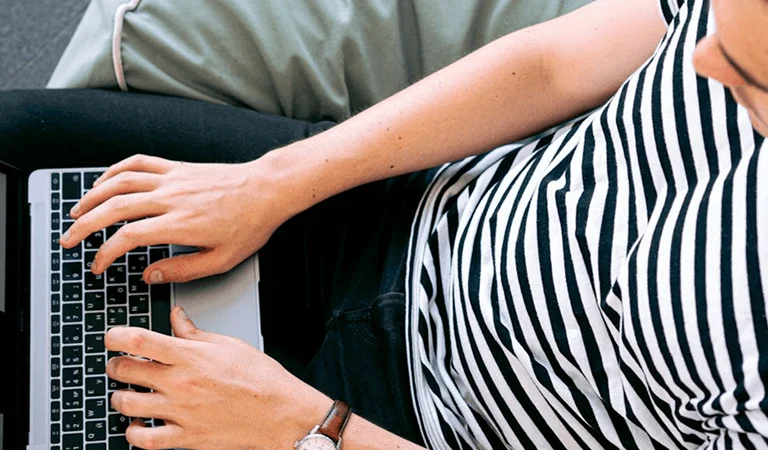 A man in a striped shirt sits on a couch, working on a laptop.