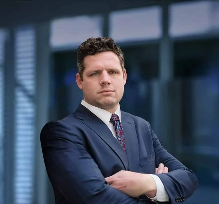xA well-dressed man stands confidently with arms crossed, displaying a professional demeanor against a blurred office background.