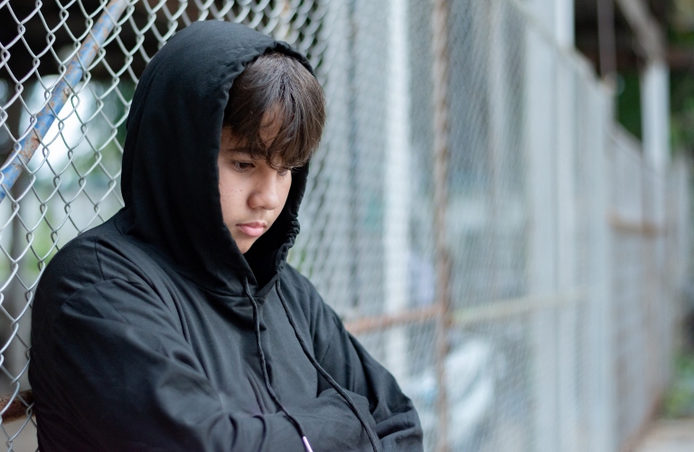 teenaged boy in a black hoodie sits with his knees pressed against a metal fence